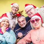 A group of five seniors, delightfully smiling and wearing festive Santa hats, gathers closely together. One dons reindeer antlers while another sports heart-shaped sunglasses, capturing the joy often found in retirement communities. They are set against a cheerful green backdrop.