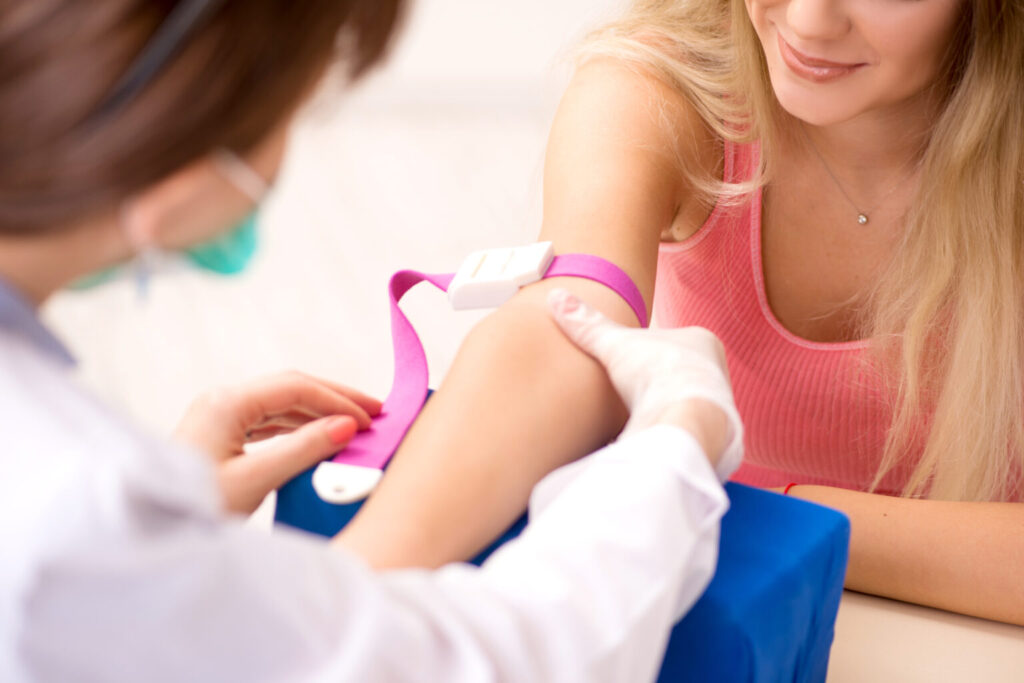 A healthcare professional, embodying the essence of a Girl Power Guide, is wearing a mask and gloves as she prepares to draw blood from a woman in her 20s, who is sitting and smiling. A pink tourniquet graces her arm against the softly blurred background, highlighting the importance of regular health checks.