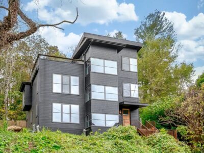 A modern black house with large windows and a wooden front door sits amid lush greenery and trees, channeling Asheville's natural allure. Its clean, angular design stands strikingly against the partly cloudy blue sky.