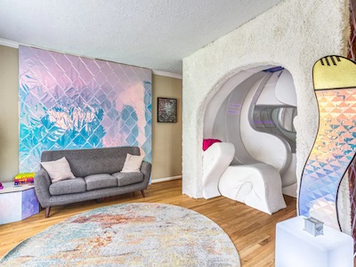 A modern, colorful living room in Portland, Oregon features a gray couch with two pillows, a round, abstract-patterned rug, and an iridescent wall panel. Embrace endless possibilities with a unique tunnel-like seating area adorned with white curved structures and a decorative surfboard.