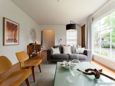 A modern living room in Portland offers endless possibilities with its gray sofa and white cushions, a glass coffee table adorned with decor, two wooden chairs, and a large window draped in curtains. Abstract art graces the walls, while a floor lamp and wooden sideboard complete the minimalist aesthetic.