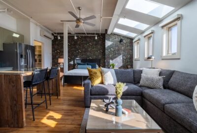 A modern loft apartment in Asheville features a gray sectional sofa with decorative pillows, a glass coffee table, and bar stools around a wooden island. In the background, there's a bed against a textured wall. Skylights illuminate the space with natural light, creating an inviting oasis perfect for relaxing or enjoying brews.