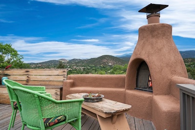 A patio with a clay fireplace, green wicker chairs, and a wooden table on a deck evokes the rich culture of Santa Fe. In the background, green trees and mountains rise under a blue New Mexico sky.