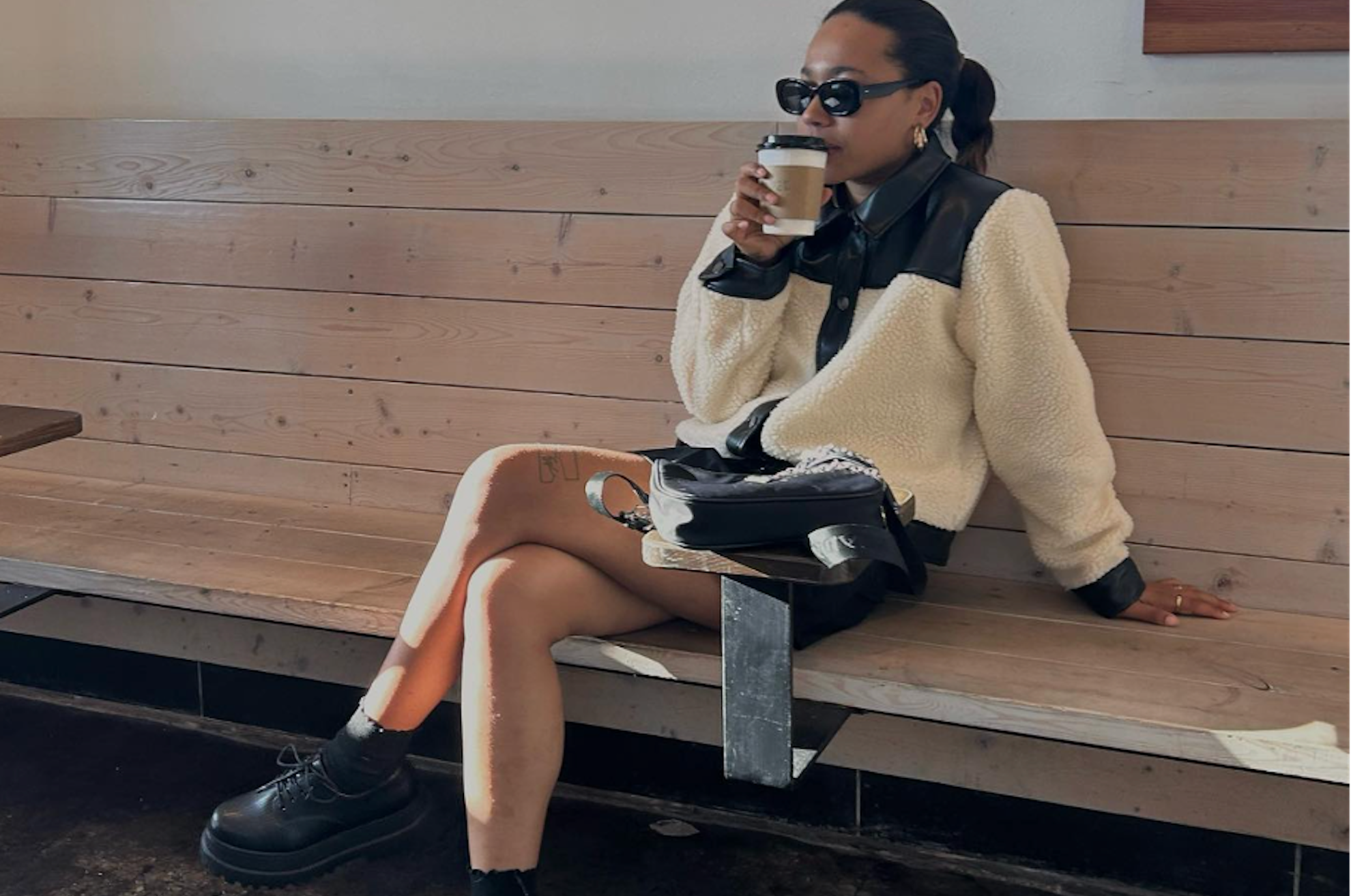 A person sits on a wooden bench in a café, embodying urban vogue with sunglasses, a cozy white and black jacket, and holding a coffee cup. They exude street style essentials with one leg crossed over the other and a chic black bag by their side.