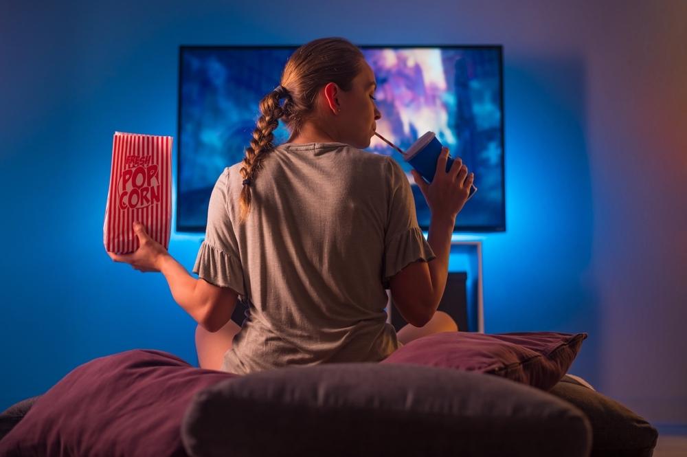 A person with a braid sits on a couch, holding popcorn and a drink, enjoying Movie Night in a dimly lit room. A blue light illuminates the scene, creating a cozy atmosphere as they dive into their Top 10 Movie Picks.