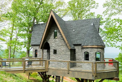 A quaint stone house nestled in the trees of Asheville, North Carolina, elevated on a wooden platform. It features an A-frame roof and a round turret, surrounded by lush greenery—a perfect retreat for sipping brews amid nature's embrace.