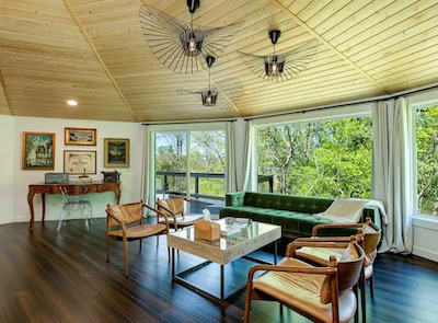 A spacious living room with a wooden ceiling and dark wood floors offers a cozy Asheville touch. It features a green velvet sofa, wooden chairs, and a glass coffee table. Large windows showcase the lush, green North Carolina view. Modern art and decor are displayed throughout for added flair.