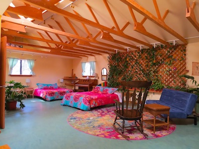 A spacious room with exposed wooden beams features two colorful beds and a blue futon. A rocking chair sits on a vibrant round rug, echoing the rich culture of Santa Fe. The back wall is adorned with a trellis covered in plants, and natural light streams in through two small windows.