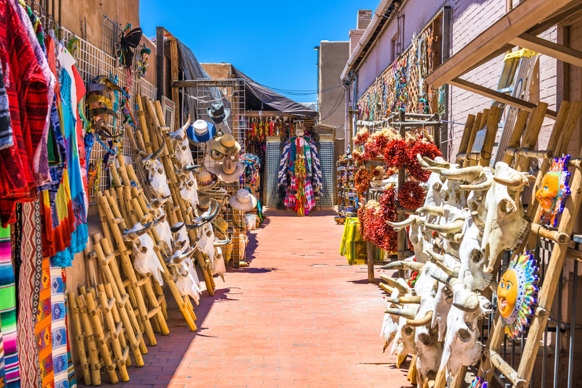 A vibrant outdoor market in Santa Fe, New Mexico, features colorful textiles, bamboo didgeridoos, decorative skulls, and assorted crafts lining a brick walkway. Brightly colored decorations hang overhead under the clear blue sky, reflecting the area's rich culture.