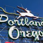 A vintage neon sign with a leaping stag proclaims "Portland, Oregon," set against a clear blue sky, embodying an aura of endless possibilities.