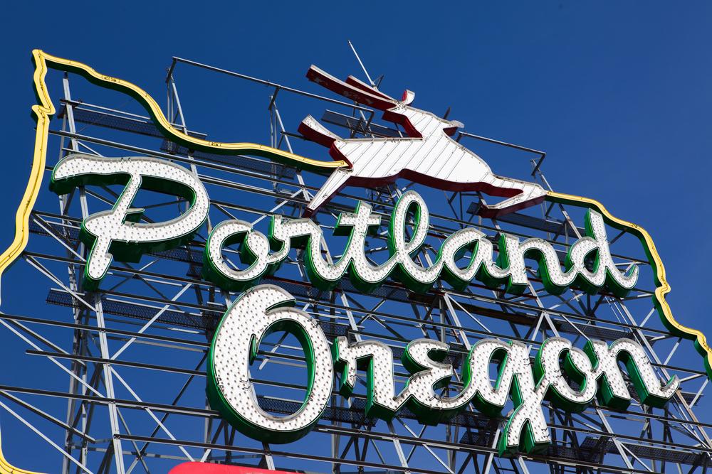 A vintage neon sign with a leaping stag proclaims "Portland, Oregon," set against a clear blue sky, embodying an aura of endless possibilities.