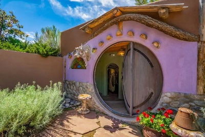 A whimsical, round door on a colorful, rustic house with purple and earthy tones evokes the rich culture of Santa Fe. The garden features lush greenery and red flowers under New Mexico's bright blue sky with clouds. A stone pathway leads to the entrance, inviting you in.