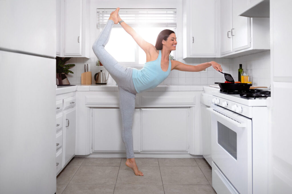 A woman in athletic wear effortlessly balances in a yoga pose, finding her zen as she cooks in a bright, modern kitchen. She extends one leg behind her and holds a spatula over the pan on the stove, surrounded by sleek white cabinets and tiled flooring.