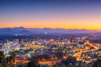 A vibrant scene unfolds over Asheville, North Carolina, at dusk with a glowing skyline and winding roads. Hills and mountains form a scenic backdrop under a colorful sunset sky, transitioning from orange to deep blue as urban lights illuminate the buildings below.