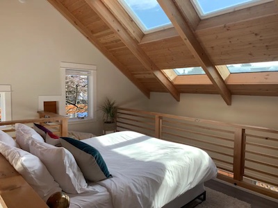 Cozy loft bedroom with wooden beams and large skylights. Sunlight fills the room, illuminating a neatly made bed with colorful cushions. A small plant near the window adds a touch of greenery, creating endless possibilities for relaxation in this serene Portland retreat.