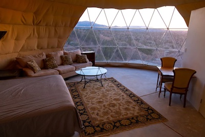 Interior of a geodesic dome with panoramic windows overlooking the enchanting desert landscape of New Mexico. The room features a bed, sofa, glass coffee table, rug, and a small table with chairs, all in a cozy Santa Fe style bathed in warm, natural light. A true cultural and outdoor escape.