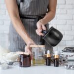 A person wearing a gray apron is preparing homemade candles. They are pouring wax from a pot into glass jars with wooden wicks. Surrounding them are jars of ingredients, dried herbs, and essential oil bottles on a white table.