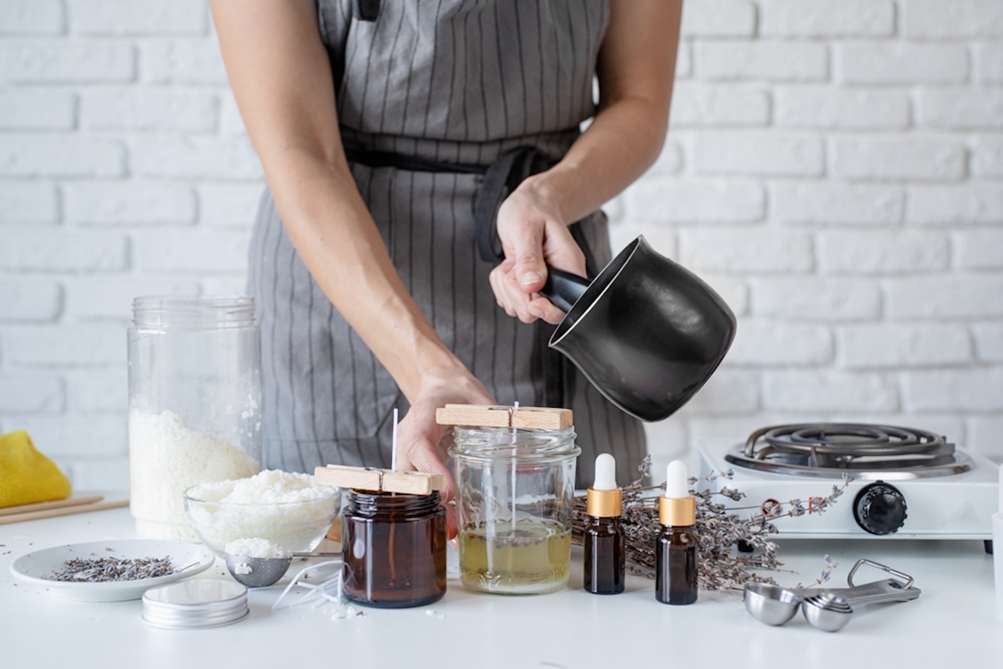 A person wearing a gray apron is preparing homemade candles. They are pouring wax from a pot into glass jars with wooden wicks. Surrounding them are jars of ingredients, dried herbs, and essential oil bottles on a white table.