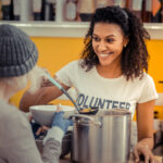 A smiling volunteer supports winter outreach efforts, serving soup to a person wearing a beanie in a community kitchen. Shelves with various items are visible in the background.