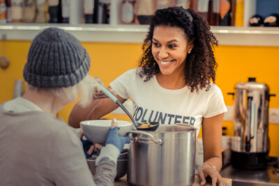 A smiling volunteer supports winter outreach efforts, serving soup to a person wearing a beanie in a community kitchen. Shelves with various items are visible in the background.