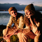 Two people sitting by a lakeside at dusk, wearing beanies and jackets. One person is playing the harmonica while the other holds a skewer. A campfire is in front of them, and city lights are visible across the water.