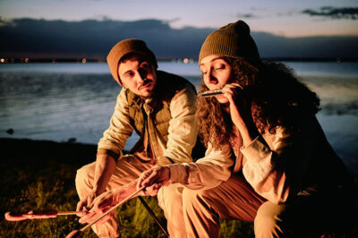 Two people sitting by a lakeside at dusk, wearing beanies and jackets. One person is playing the harmonica while the other holds a skewer. A campfire is in front of them, and city lights are visible across the water.