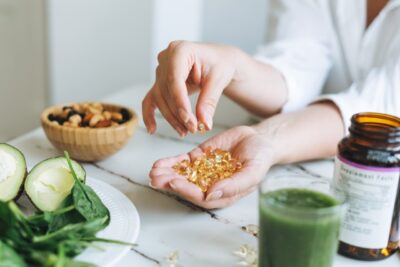 A person holds fish oil capsules over a table adorned with fresh spinach, avocado halves, mixed nuts, and a green smoothie. An amber glass supplement bottle suggests a focus on vivacious vitality and nutrition.