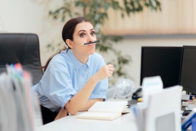 A woman in a blue shirt is sitting at a desk, playfully balancing a pen on her upper lip, perhaps trying to overcome procrastination. Her elbow rests on an open notebook as monitors and office supplies surround her, suggesting she's breaking free from distractions.