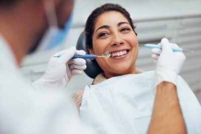 A woman in a dental chair smiles with relief as the dentist, offering tips to ease dentist fear, examines her teeth. She wears a bib while the dentist holds tools. The setting is a serene dental clinic, with both appearing positive and focused.