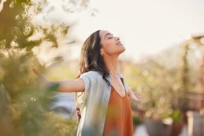 A woman with long hair stands outdoors, arms outstretched, savoring the sunlight. Wearing a light shirt over an orange top, she embraces this extraordinary day. The softly blurred greenery in the background enhances the serene atmosphere, turning mundane moments into something special.