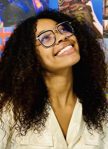 A person with curly hair and glasses smiling upwards against a colorful background. They are wearing a light-colored top.