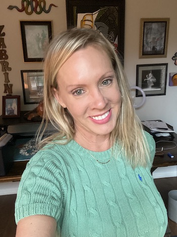 A woman with blonde hair smiles while taking a selfie indoors. She is wearing a green cable-knit sweater, and there are various framed artworks and decorations on the wall behind her.