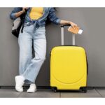 A person stands against a gray wall holding a passport and boarding pass. They wear a yellow top and blue jeans, with a denim jacket tied at the waist. A yellow suitcase stands beside them, and they have a backpack over one shoulder.