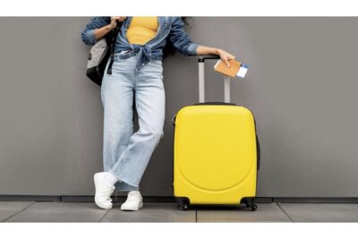 A person stands against a gray wall holding a passport and boarding pass. They wear a yellow top and blue jeans, with a denim jacket tied at the waist. A yellow suitcase stands beside them, and they have a backpack over one shoulder.