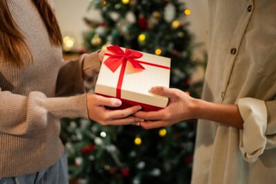 Two people exchanging a gift wrapped in beige paper with a red ribbon, in front of a decorated Christmas tree with lights and ornaments. One person wears a beige sweater, and the other wears a light-colored shirt.