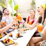 A group of friends savors a budget-friendly outdoor brunch, laughing and clinking glasses filled with colorful drinks. Plates decorated with fruit adorn the table. Everyone is smiling, wearing casual summer clothes, surrounded by greenery—a perfect example of savvy hosting tips in action.