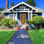 A charming blue house with white trim, a front porch, and a red door offers standout curb appeal. The neatly landscaped yard features green grass, bushes, and two large red planters by the entrance. A concrete path leads to this wow factor porch.