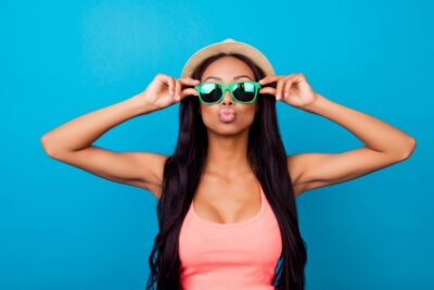 Woman wearing a hat and green sunglasses poses playfully against a blue background, making a kissy face and holding her sunglasses with both hands—a perfect snapshot of Spring Break essentials. She has long dark hair and wears a pink tank top.